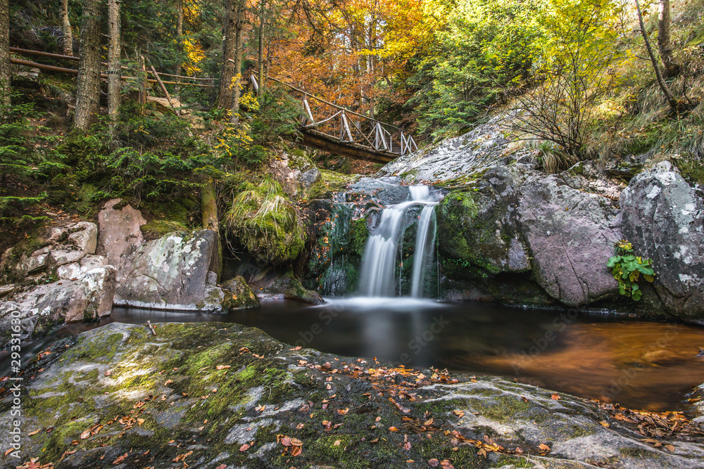 Small creek waterfall in a beautiful deciduous autumn forest. Bright autumn leaves on stones covered with moss by the river. Beautiful autumn landscape with wooden bridge, Long exposure.