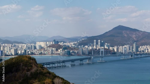 Timelapse modern Busan city parts with skyscrapers connected by bridge over ocean bay against green hillpeak with shadows from floating in blue sky clouds zoom in photo