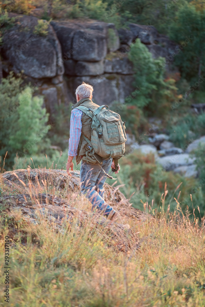 Back view of tourist man with a gray beard with a backpack on his shoulders against the backdrop of the gorge, rocks and stones, the concept of tourism and outdoor activities in old age