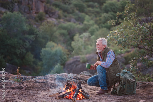 Eldery tourist with gray hair and beard sits on the edge of a canyon near a burning fire with a mug in his hands, against the backdrop of the sun, mountains and trees