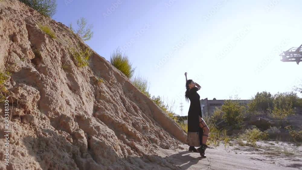 Sexy asian girl in a long black dress and heavy boots walking on the background of desert sand. Go-go show at techno festival. The concept of post-apocalyptic wasteland. Steady around Sexy asian girl in a long black dress and heavy boots walking on the background of desert sand. Go-go show at techno festival. The concept of post-apocalyptic wasteland. Steady around