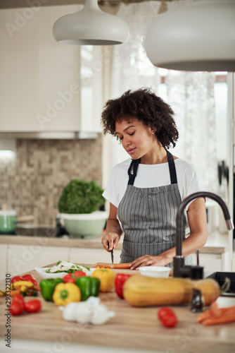 Attractive mixed race woman in apron cutting carrot and preparing dinner while standing in kitchen. On kitchen counter are vegetables.