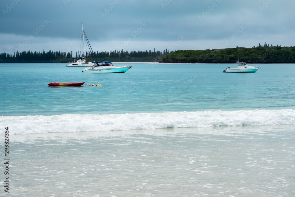 Boats on the water and waves crashing on the beach at Kuto Bay with pine forests at the horizon in the background on a beautiful day at Isle of Pines in New Caledonia, South Pacific Ocean.