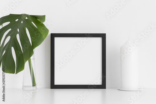Black square frame mockup with a monstera leaf in a glass vase and a candle  on a white table.