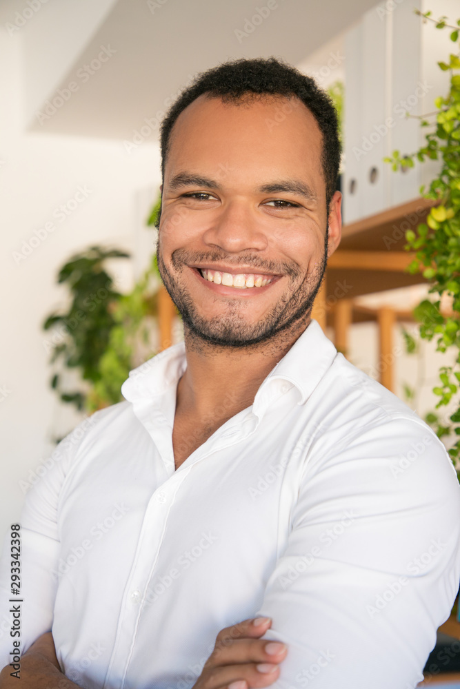 Happy cheerful male employee in casual posing in modern office. Handsome young man in white shirt, standing with arms folded and smiling at camera. Male portrait concept
