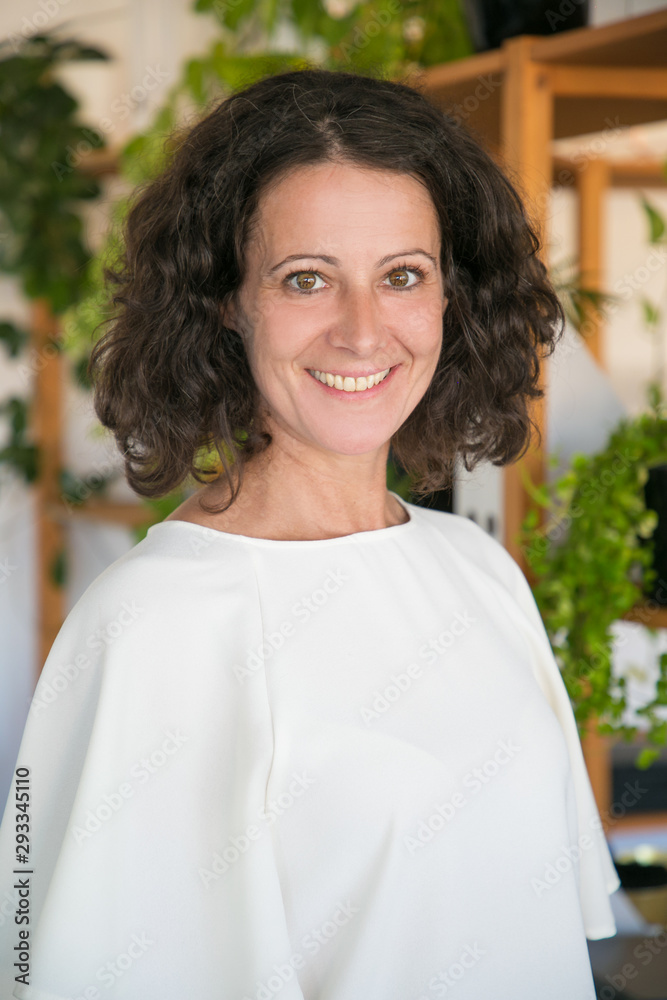 Happy confident employee in casual posing in modern office. Middle aged woman with curly hair smiling at camera. Female portrait concept Happy confident employee in casual posing in modern office. Middle aged woman with curly hair smiling at camera. Female portrait concept