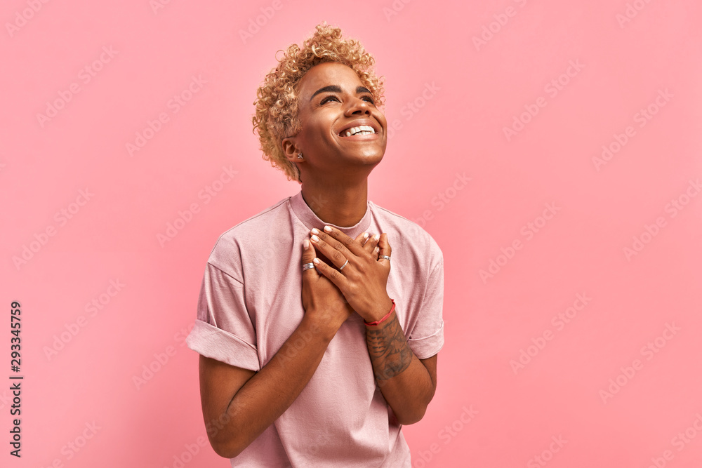 Touched beautiful smiling Afro American girl keeps palms on heart, expresses appreciation, pleasure and gratitude, wears a lavender t-shirt, models over pink background. Confession in love concept - obrazy, fototapety, plakaty 