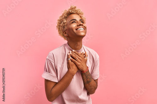Touched beautiful smiling Afro American girl keeps palms on heart, expresses appreciation, pleasure and gratitude, wears a lavender t-shirt, models over pink background. Confession in love concept photo