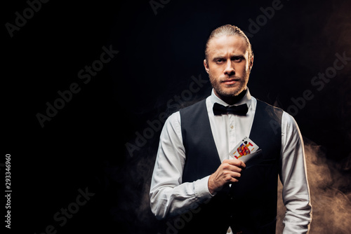 Foto handsome bearded croupier holding playing cards on black with smoke