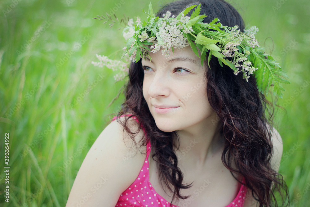 a young dark haired girl in a wreath of wild flowers and a pink sundress sits in a field and smiled