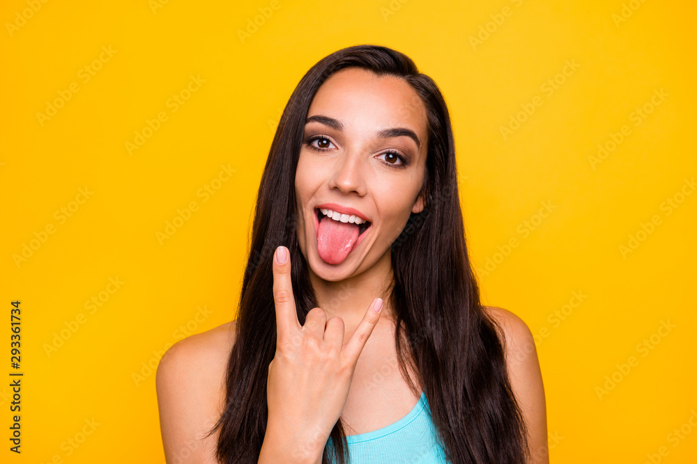 Close up photo of playful girlish feminine young rocker sticking tongue out with fingers horned showing her love for metal isolated over vivid color yellow background