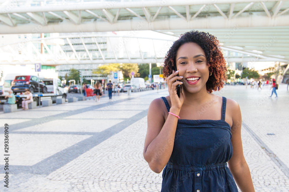 Cheerful excited woman talking on cell while walking down city street. Young black woman going outside, speaking on mobile phone, smiling. Phone talk concept