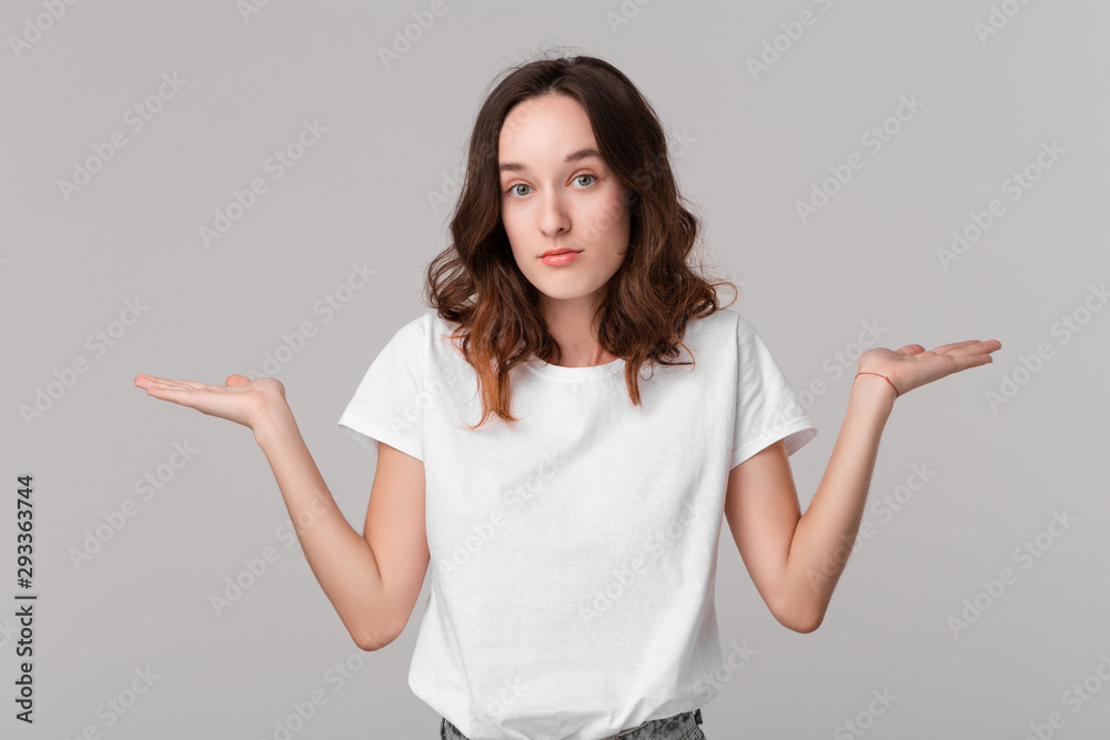 Pretty brunette woman in a white tee shrugging her shoulders beacause she is indignant standing isolated over grey background. Copy space of advertising. Pretty brunette woman in a white tee shrugging her shoulders beacause she is indignant standing isolated over grey background. Copy space of advertising.