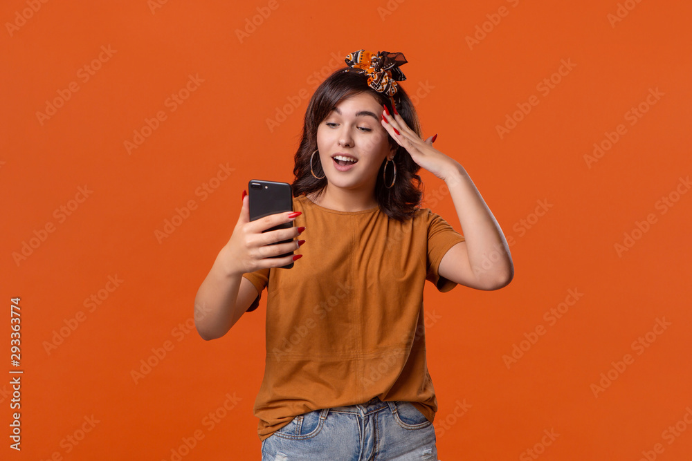 Pretty brunette woman in a t-shirt and beautiful headband checking smartphone for notifications and feeling excited standing isolated over orange background. Daily communication Pretty brunette woman in a t-shirt and beautiful headband checking smartphone for notifications and feeling excited standing isolated over orange background. Daily communication