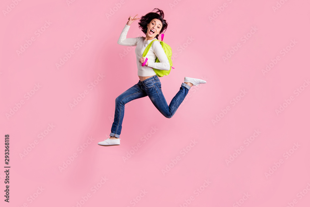 Full body photo of lady jumping high holding green rucksack running to shopping showing v-sign wear pullover and jeans isolated pink color background