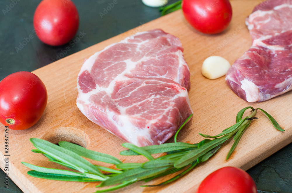Two raw juicy slices of pork, steaks with red tomatoes, rosemary and garlic on a cutting board close-up, top view Two raw juicy slices of pork, steaks with red tomatoes, rosemary and garlic on a cutting board close-up, top view