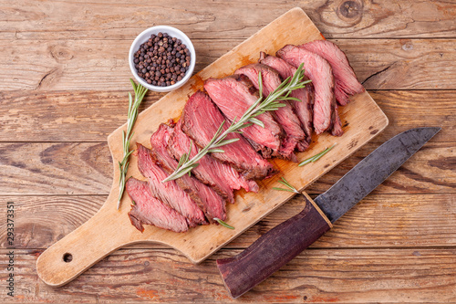 Slices of medium rare roast beef meat on wooden cutting board, hunters knife, pepper and rosemary twigs on wooden background. Gourmet food. Raw meat beef steak. Top view.