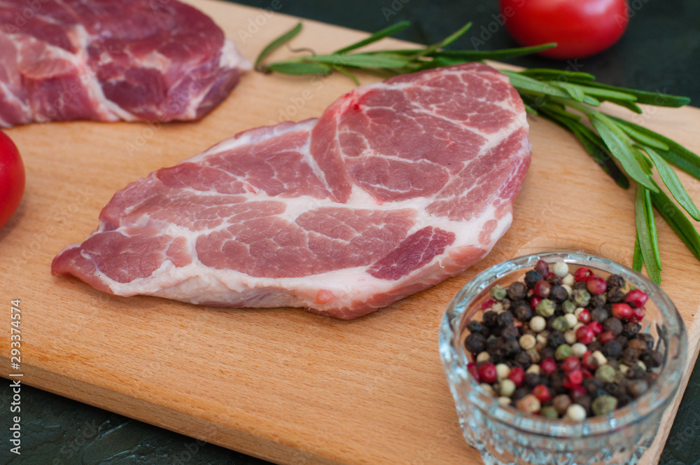 Raw pork steaks, rosemary, tomatoes and peppers on wooden cutting board, dark background, side view