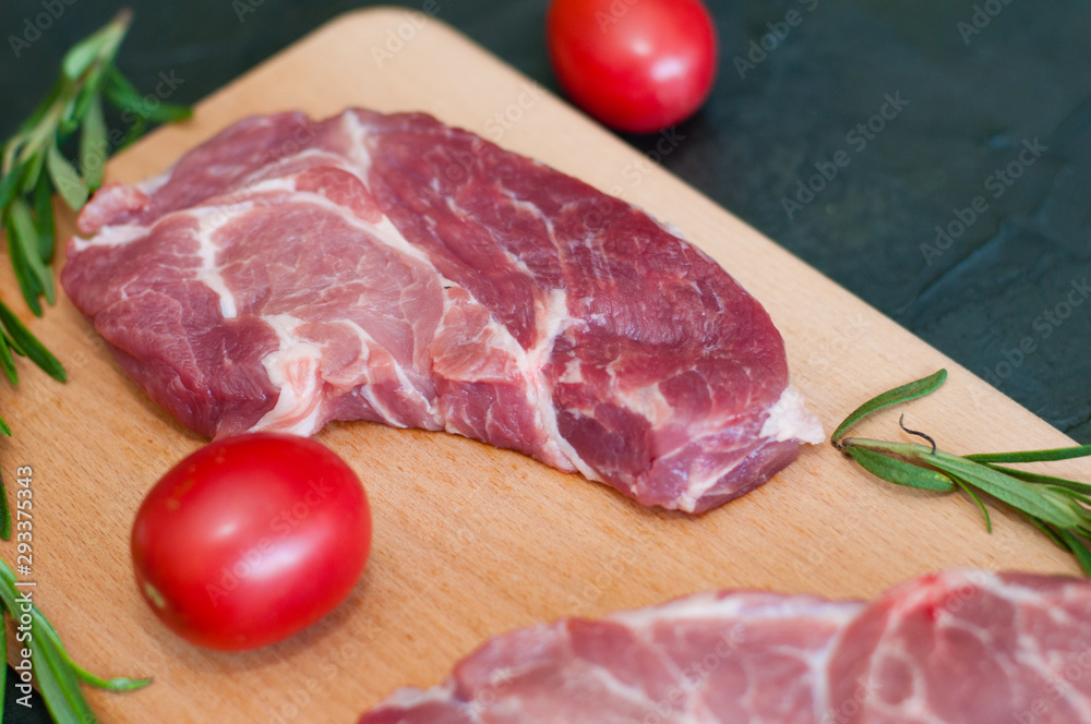 Raw pieces of pork, rosemary and tomatoes on a cutting board, on dark gray concrete background, close-up Raw pieces of pork, rosemary and tomatoes on a cutting board, on dark gray concrete background, close-up