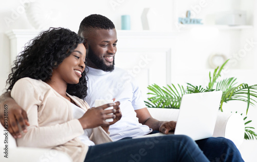 Cheerful black couple sitting on couch looking at laptop screen