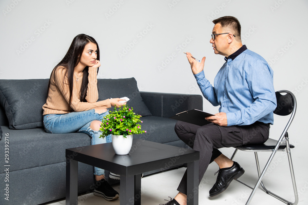 Beautiful young woman in casual clothes is sitting on the couch at the psychotherapist. Handsome middle aged doctor is holding a folder and talking to her Beautiful young woman in casual clothes is sitting on the couch at the psychotherapist. Handsome middle aged doctor is holding a folder and talking to her