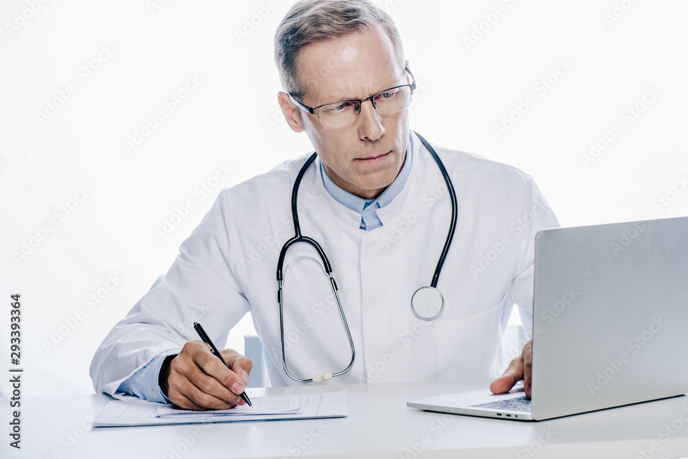 handsome doctor in white coat writing and using laptop isolated on white handsome doctor in white coat writing and using laptop isolated on white