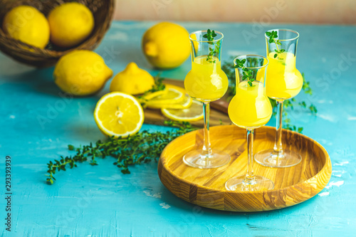 Limoncello with thyme in three grappas wineglass in wooden tray, fresh lemon in basket on light concrete table. Artistic still life on light background photo