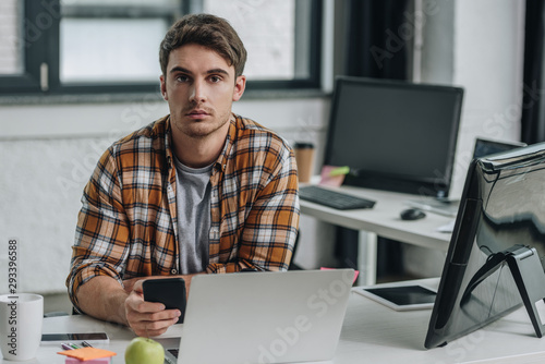serious young programmer looking at camera while sitting at workplace and holding smartphone