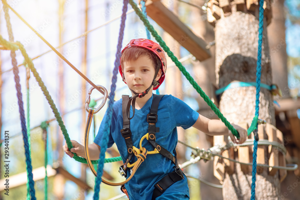 Boy enjoying activity in climbing adventure park at sunny summer day. Kid climbing in rope playground structure. Safe climbing with helmet insurance. Child in forest adventure park, extreme sport. Boy enjoying activity in climbing adventure park at sunny summer day. Kid climbing in rope playground structure. Safe climbing with helmet insurance. Child in forest adventure park, extreme sport.