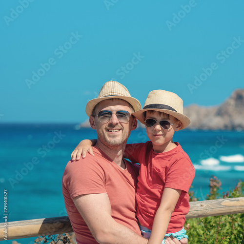 Cute European boy is on his father’s hands in front of picturesque wavy sea, enjoying summer holidays. They wearing similar clothes sun hats and sunglasses smiling and looking towards camera.