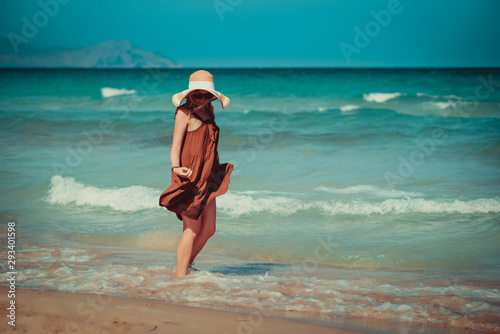 Young European lady in colorful bonnet hat and beautiful dress walking along picturesque wavy sea, she enjoying serenity around. photo