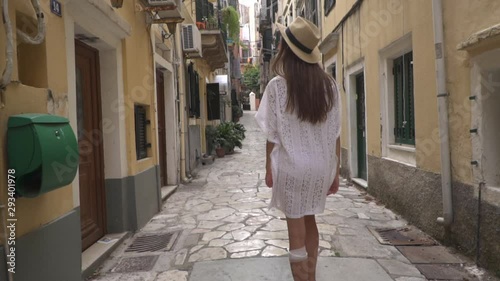 Rear view beautiful girl in white dress walking narrow european streets in old town, Kerkyra, Greek Island Corfu. Summer travel vacation. Adventure.