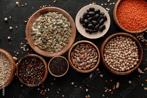 top view of wooden bowls and plates with raw assorted beans, cereals and seeds on dark surface with scattered grains