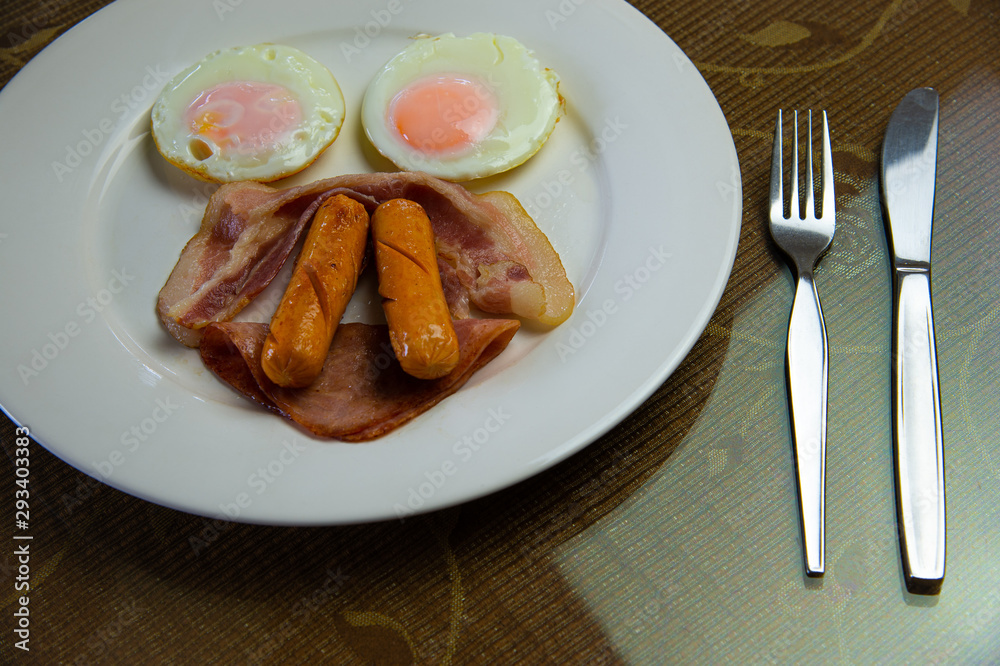 Yummy American breakfast with sunny fried eggs, rosted bacon, ham, two sausages in white dish with silver fork and knife on table. Yummy American breakfast with sunny fried eggs, rosted bacon, ham, two sausages in white dish with silver fork and knife on table.