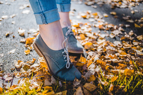 Women in cadet blue leather boots standing on yellow autumn birch leaves on asphalt. Her jeans are tucked up.