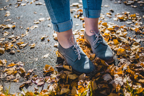 Women in cadet blue leather boots standing on yellow autumn birch leaves on asphalt. Her jeans are tucked up.
