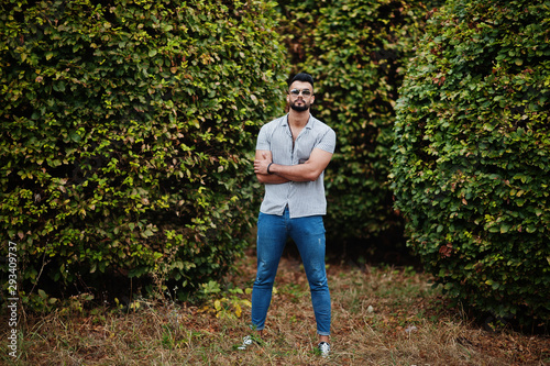 Fashionable tall arab beard man wear on shirt, jeans and sunglasses posed on park against greenery.
