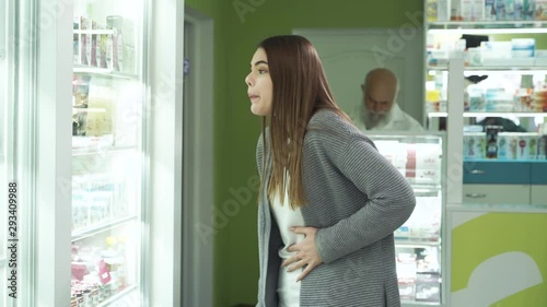 Young caucasian brunette girl with severe pain in the stomack staying at the pharmacy and choosing the pain medication. Senior pharmacist on the background waiting for his customer. Healthcare. photo