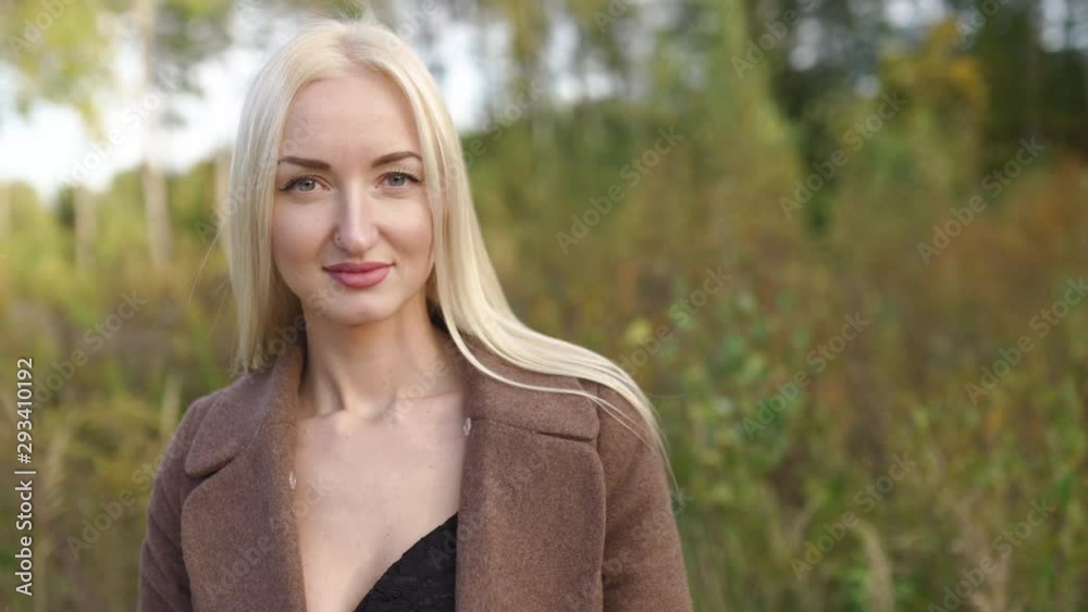 Blonde woman portrait in outside autumn park with golden and green forest in the background - Wearing a brown coat Blonde woman portrait in outside autumn park with golden and green forest in the background - Wearing a brown coat