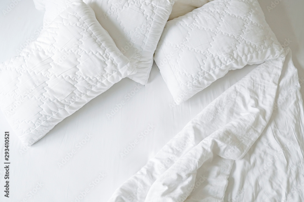 Obraz premium Unmade hotel room bed with bunch of puffy pillows in pile, perfectly clean snow white sheets and blanket in natural sun light. Close up, copy space for text.