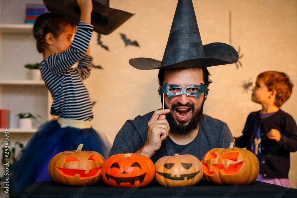 Fototapeta premium Happy family: father, brother and sister celebrate Halloween. Cheerful children in carnival costumes indoors. Cheerful children and parents play with pumpkins and black witch hats