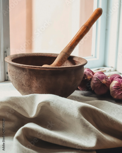 rustic flat lay, clay mortar with wooden pestle for grinding spices on linen background and string of onions on windowsill