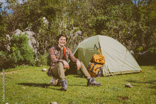 Young camper man is sitting on the rock and smiling to the camera in front of his tent. Young camper man is smiling near his tent, backpack and batons in front of the trees on the green grass.