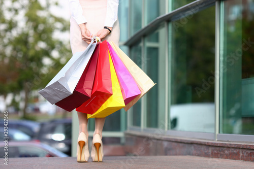 Focus on beautiful model in light clothes standing near fashionable boutique and holding multicolored shop packages. Fashion and shopping concept. Blurred background