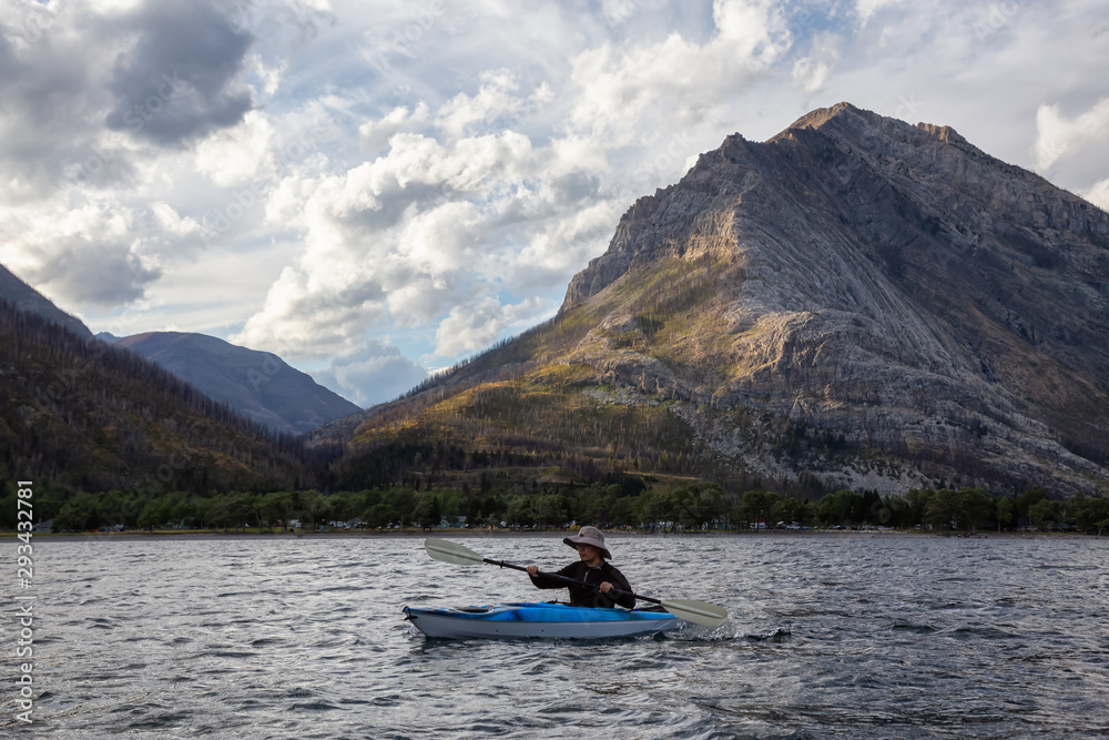 Adventurous Man Kayaking in Glacier Lake surrounded by the beautiful Canadian Rocky Mountains during a cloudy summer sunset. Taken in Upper Waterton Lake, Alberta, Canada.