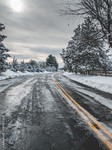 A portrait, vertical shot of a road with snow and ice after a harsh winter storm. The bright yellow lines show through in various spots. The sun peaks through the dark clouds on this country road.