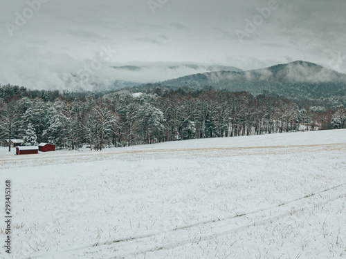 A landscape, horizontal frame of a country field and red home after a severe winter storm. Storm clouds break up over the mountains displaying beautiful snow covered fields, trees, and mountains.