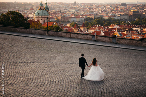 Photography Newlyweds walk on wide square of cobblestones, which offers views of city