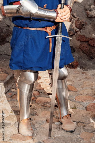 Medieval knight holding a sword in his hands. stands in front of a stone wall. lower body.