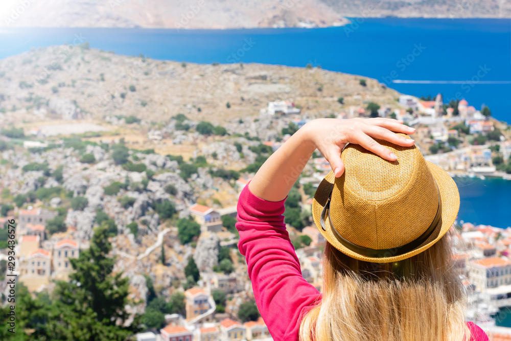 Young blonde girl with hair is sitting on top of the mountain and watching down on tiny colorful houses and Mediterranean sea on greek island Symi in Dodekanisos archipelago in bright sunbeams